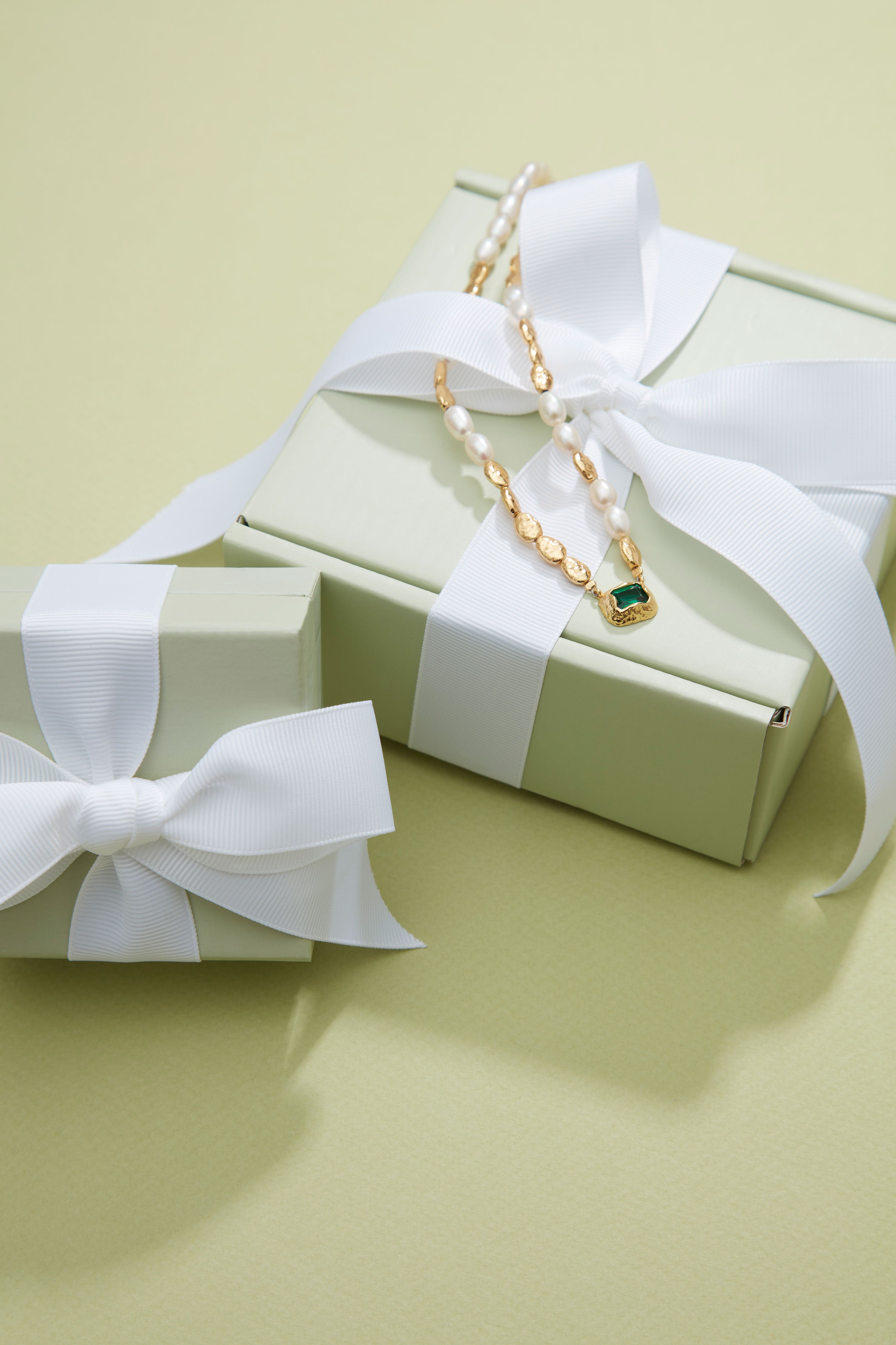Necklace with a green gemstone on a white ribbon tied around a gift box on a light green background