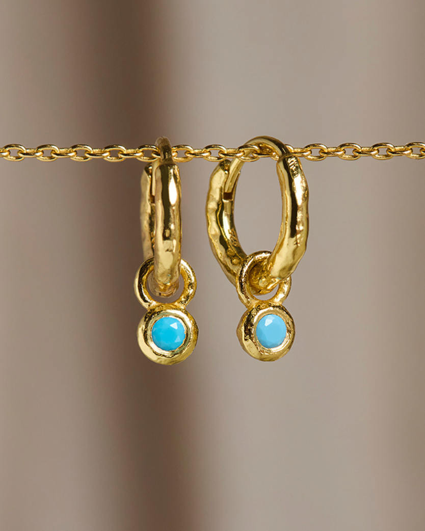 A pair of gold huggie hoop earrings with small round light blue turquoise-like drops, hanging from a gold chain against a soft beige background.