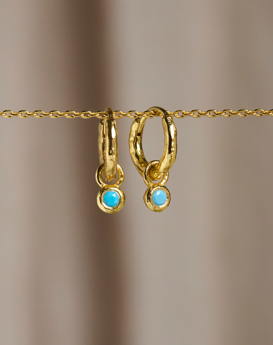 A pair of gold huggie hoop earrings with small round light blue turquoise-like drops, hanging from a gold chain against a soft beige background.