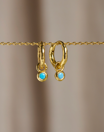 A pair of gold huggie hoop earrings with small round light blue turquoise-like drops, hanging from a gold chain against a soft beige background.